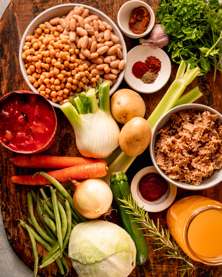 An overhead shot shows ingredients needed for a vegan minestrone soup against a rough wooden background.
