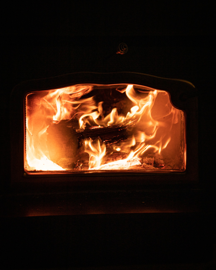 A head-on shot shows the window of a wood burning stove with a roaring fire inside. The surrounding area is completely dark.