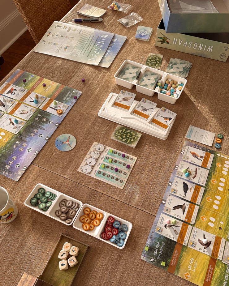 An overhead shot shows the board game Wingspan spread out on a dining room table in natural, soft sunlight.