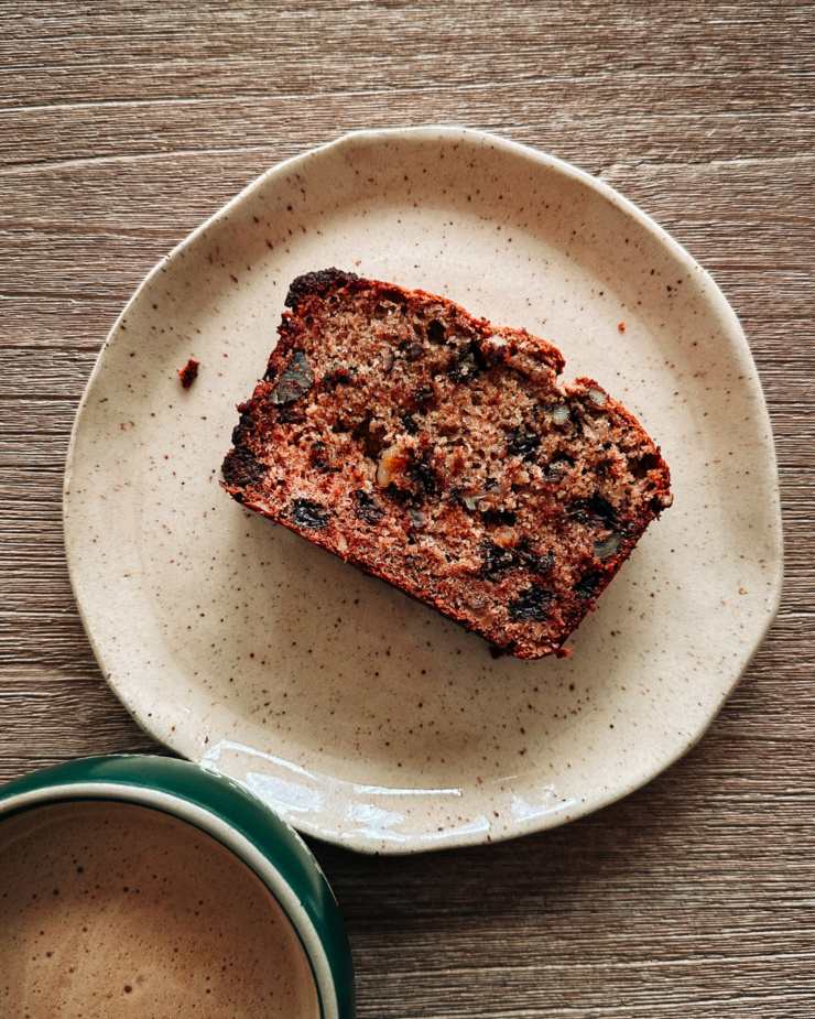 An overhead shot shows a slice of banana bread on a plate and a mug of hot coffee is nearby.