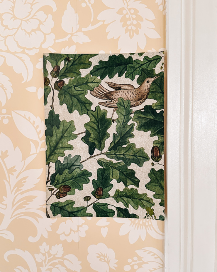 A head-on shot shows a sample of wallpaper taped on a wall. The wallpaper features oak leaves, acorns, and mourning doves.