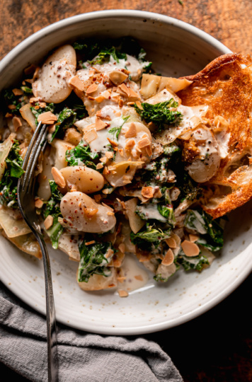 An up close, overhead shot shows an individual serving of sautéed cabbage, kale & butter beans with mustard cream. A crust of toasted bread is sticking out of the serving in a wide ceramic bowl. A fork is sticking out of the bowl also. The dish is garnished with chopped toasted almonds.