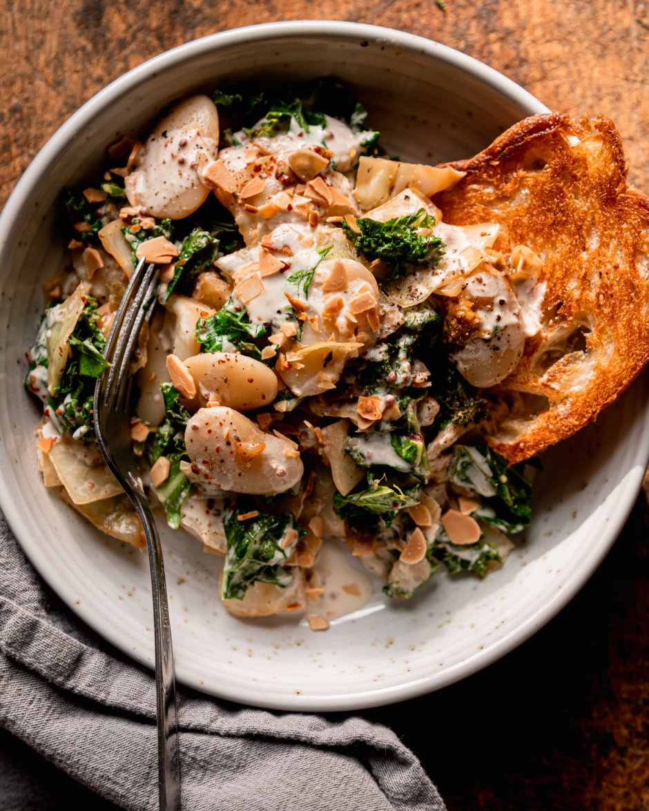 An up close, overhead shot shows an individual serving of sautéed cabbage, kale & butter beans with mustard cream. A crust of toasted bread is sticking out of the serving in a wide ceramic bowl. A fork is sticking out of the bowl also. The dish is garnished with chopped toasted almonds.