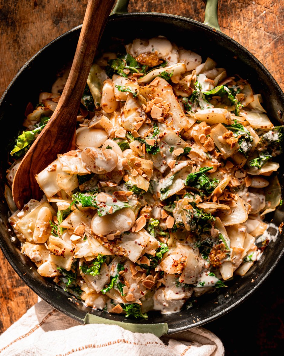 An overhead shot shows a skillet filled with sautéed cabbage, kale, and butter beans. Everything has a nice coating of vegan mustard cream and is garnished with chopped toasted almonds.