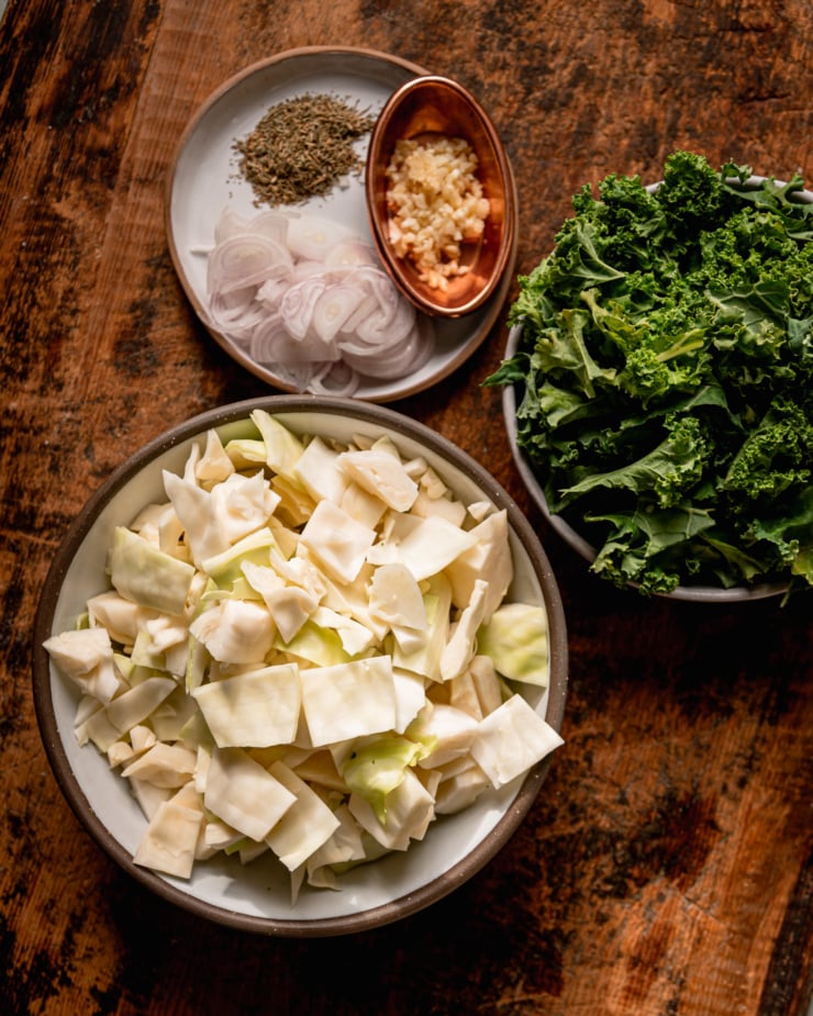 An overhead shot shows minced garlic in a small dish, dried thyme, sliced shallots, chopped kale, and chopped cabbage.