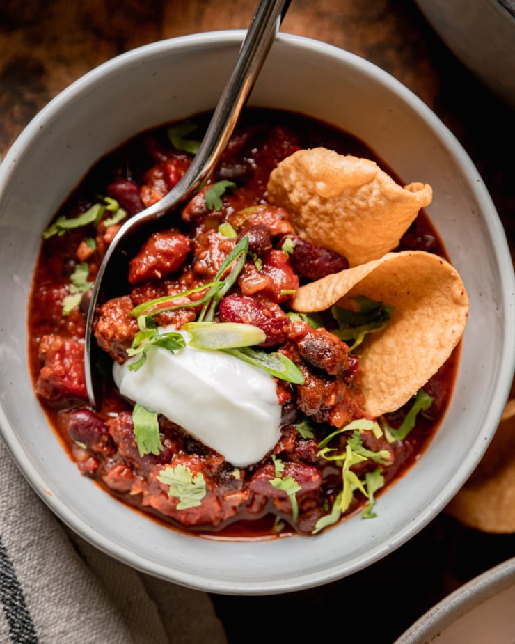 An overhead shot shows a bowl of vegan chili garnished with vegan sour cream, chopped cilantro, green onions, and a couple of tortilla chips stuck into the top. A spoon is sticking out of the bowl.