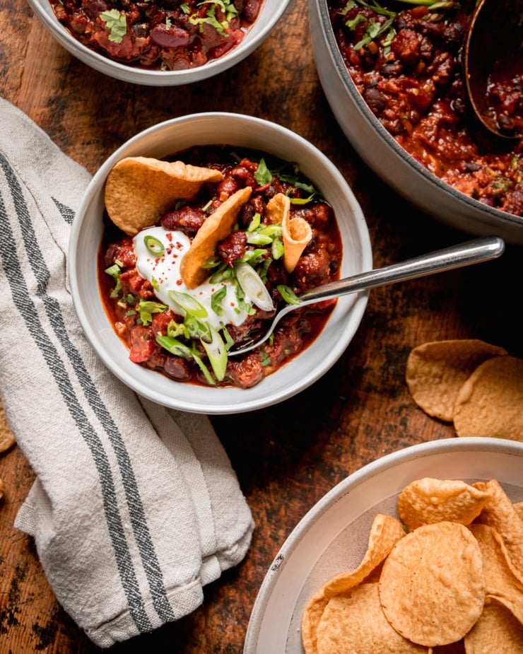 An overhead shot shows an individual serving bowl of vegan chili garnished with vegan sour cream and tortilla chips. The pot of chili and a bowl of the tortilla chips are nearby.