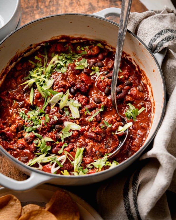 A 3/4 angle shot shows a Dutch oven-style pot filled with vegan chili that features black beans, red kidney beans, and cooked bits of vegan "sausages." The chili is garnished with chopped green onions and cilantro.