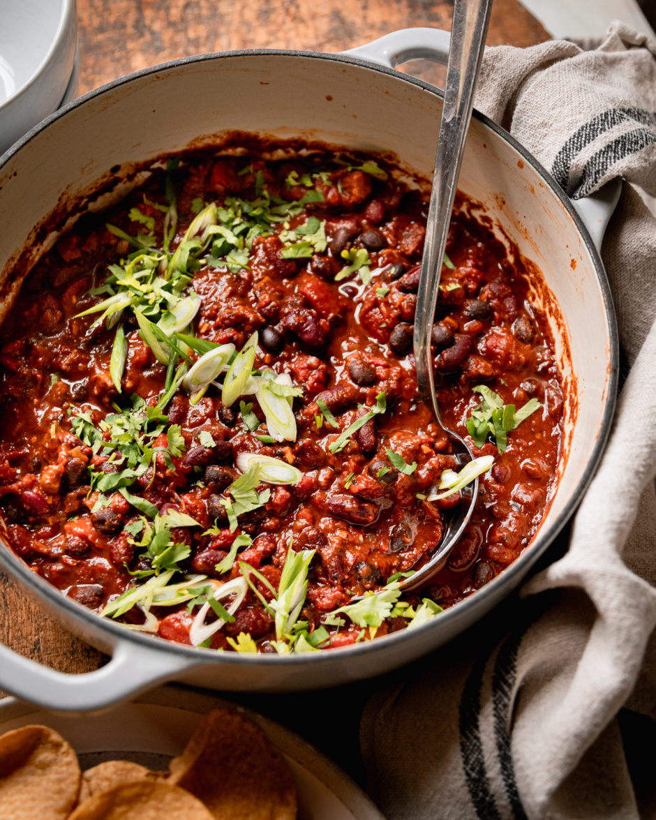 A 3/4 angle shot shows a Dutch oven-style pot filled with vegan chili that features black beans, red kidney beans, and cooked bits of vegan "sausages." The chili is garnished with chopped green onions and cilantro.