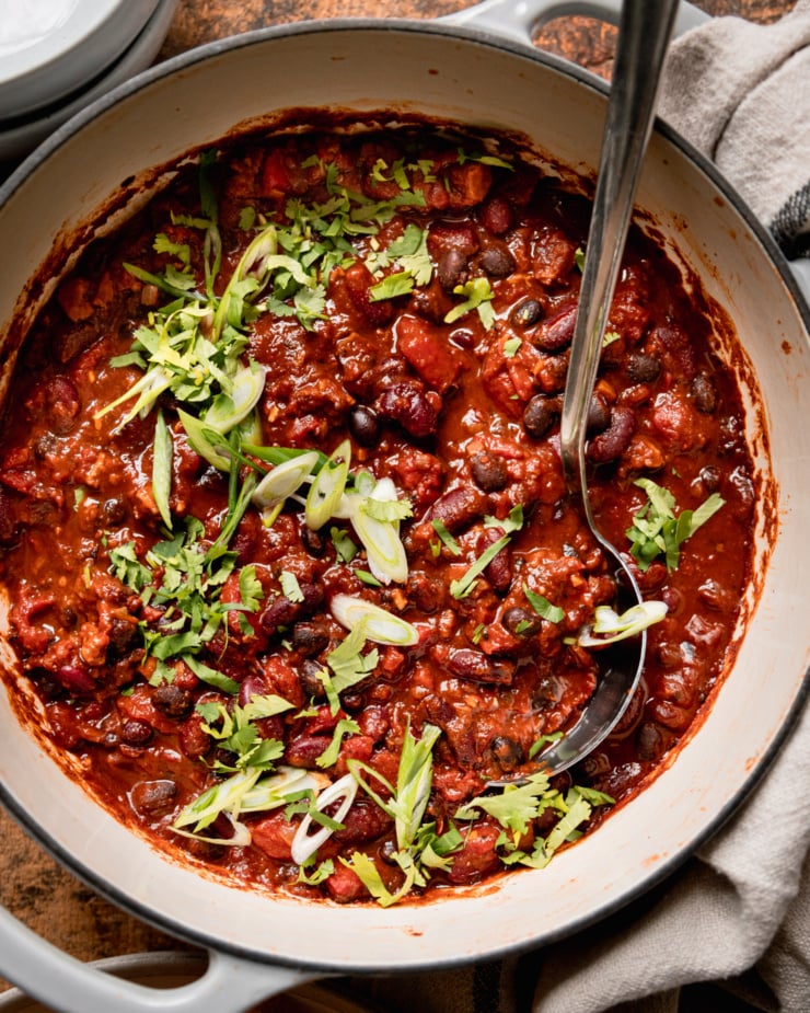 An up close, overhead shot shows a Dutch oven-style pot filled with vegan chili that features black beans, red kidney beans, and cooked bits of vegan "sausages." The chili is garnished with chopped green onions and cilantro.