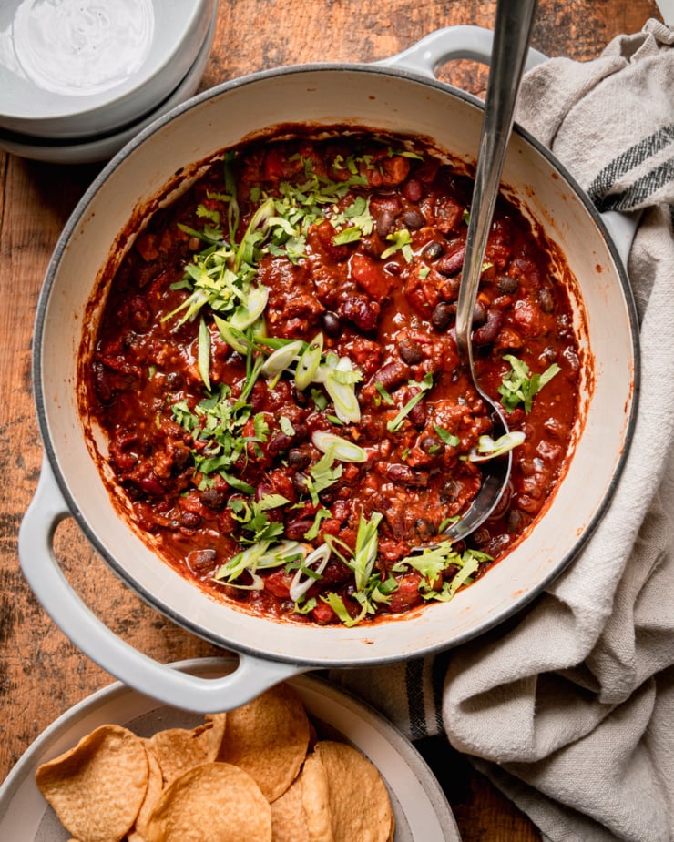 An overhead shot shows a Dutch oven-style pot filled with vegan chili that features black beans, red kidney beans, and cooked bits of vegan "sausages." The chili is garnished with chopped green onions and cilantro.