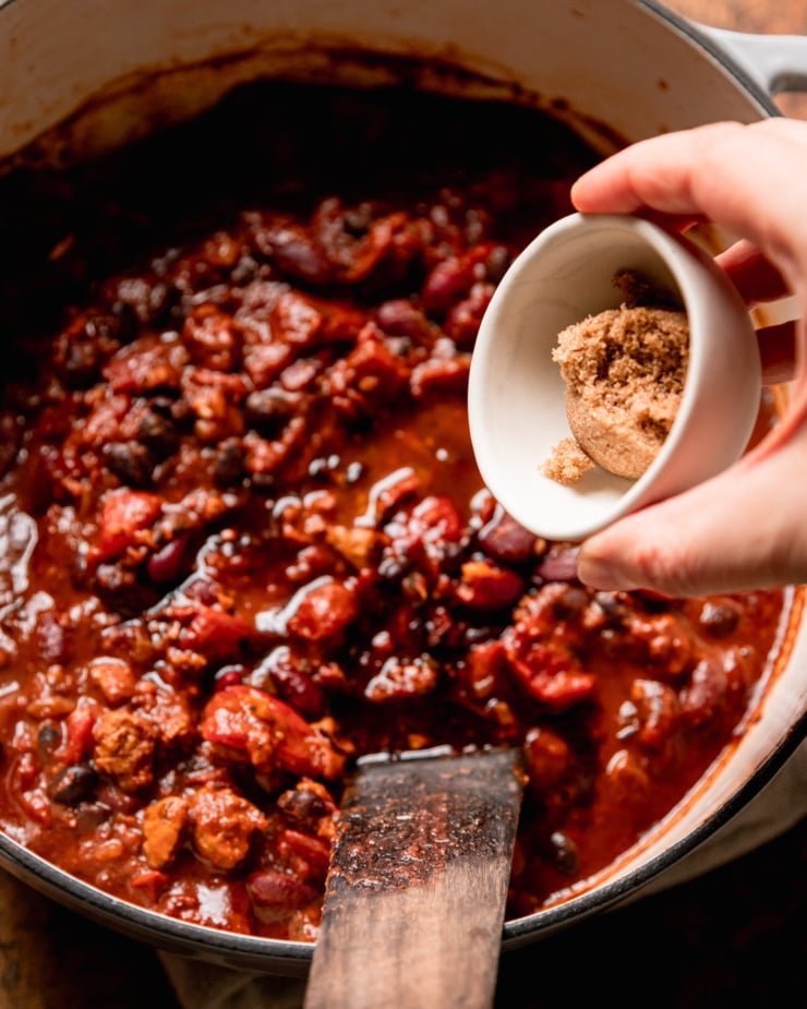An overhead shot shows a hand emptying a small bowl with brown sugar in it into a pot of chili.