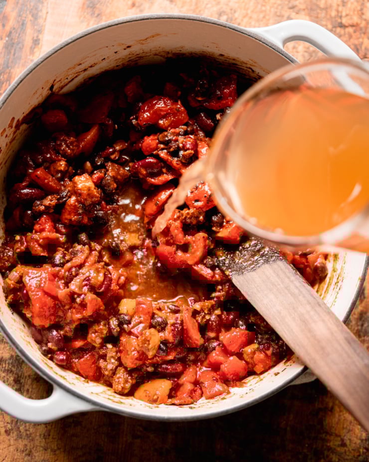 An overhead shows vegetable stock being added to a pot with diced tomatoes and beans.