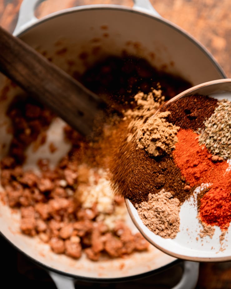 An overhead shot shows a plate of different spices being shaken into a pot with sautéed veggie sausage bits and onions.