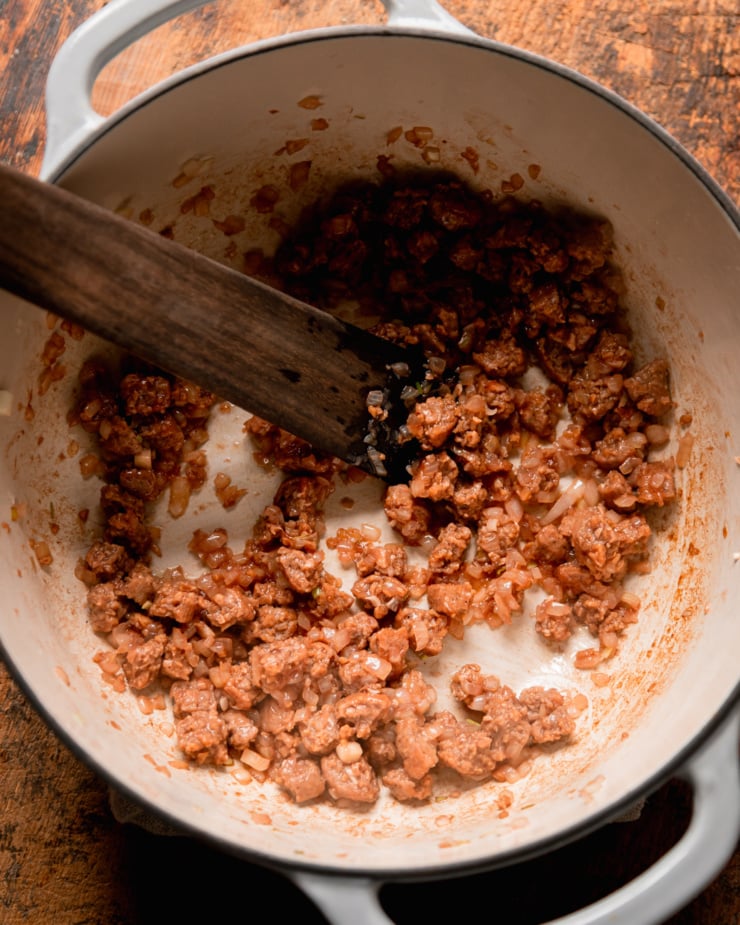 An overhead shot shows a Dutch oven with sautéed onions and vegan "sausage" bits. A wooden utensil is sticking out of the pot.