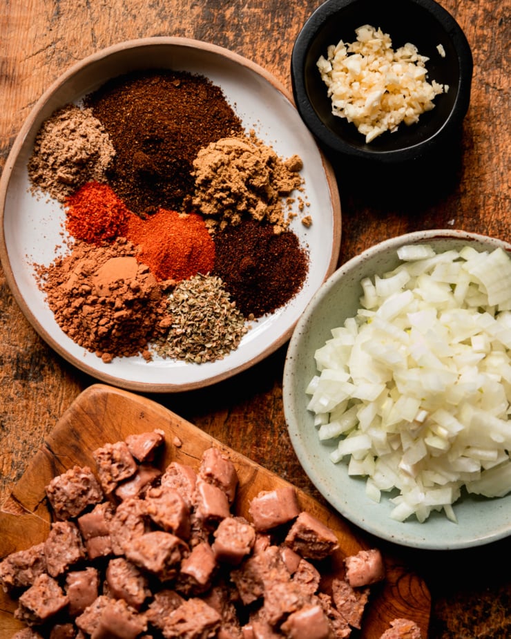 An overhead shot shows minced garlic in a small bowl, a plate with little piles of 8 different spices, a bowl with diced onions, and a small cutting board with chopped up vegan "sausages."