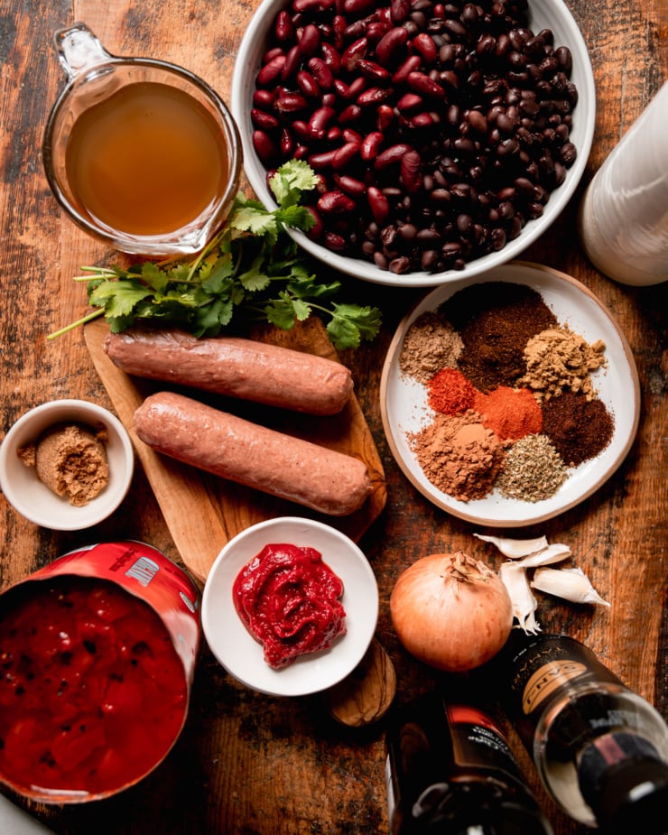 An overhead shot shows ingredients laid out: red kidney and black beans, avocado oil, a plate with different spices, garlic cloves, an onion, Tamari, apple cider vinegar, tomato paste, fire roasted diced tomatoes, vegan :sausages," brown sugar, cilantro, and vegetable stock. All are captured on a worn wooden board.