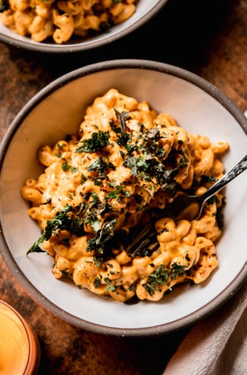 An overhead shot shows a bowl of creamy truffle mac pasta topped with crushed up bits of crunchy baked kale. A fork is sticking out of the individual serving. A lit candle is nearby.
