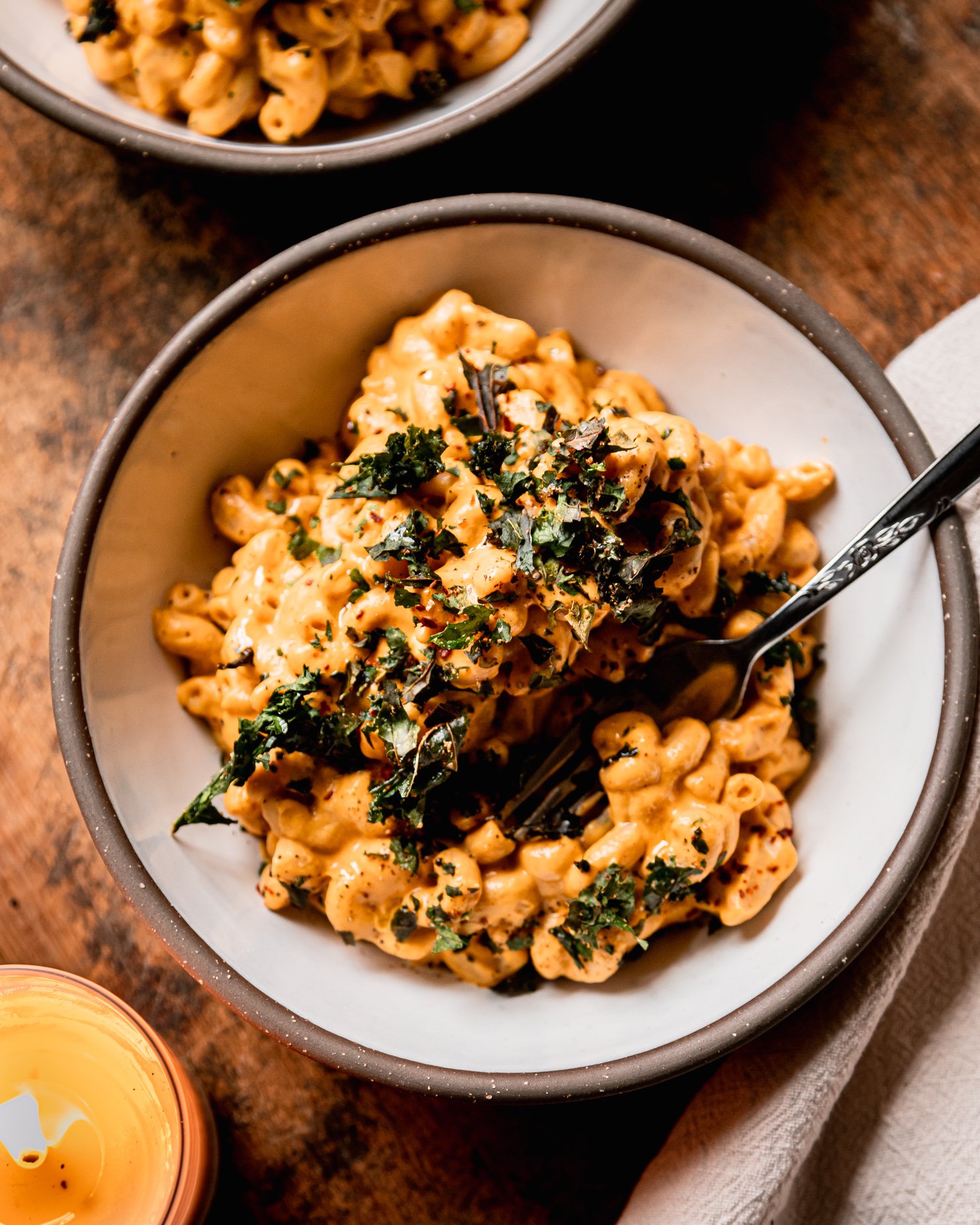 An overhead shot shows a bowl of creamy truffle mac pasta topped with crushed up bits of crunchy baked kale. A fork is sticking out of the individual serving. A lit candle is nearby.