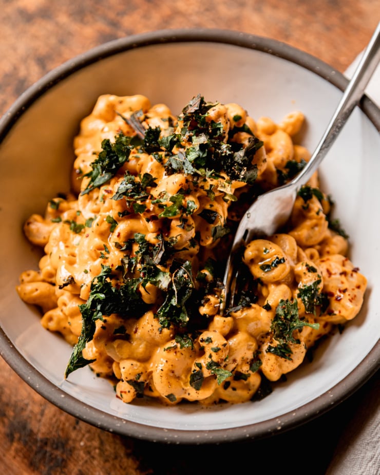 An up close, slight 3/4 angle shot shows a bowl of creamy truffle mac pasta topped with crushed up bits of crunchy baked kale. A fork is sticking out of the individual serving.