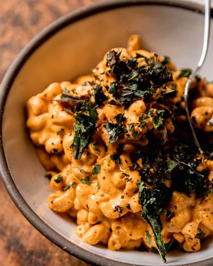 An up close, slight 3/4 angle shot shows a bowl of creamy truffle mac pasta topped with crushed up bits of crunchy baked kale. A fork is sticking out of the individual serving.