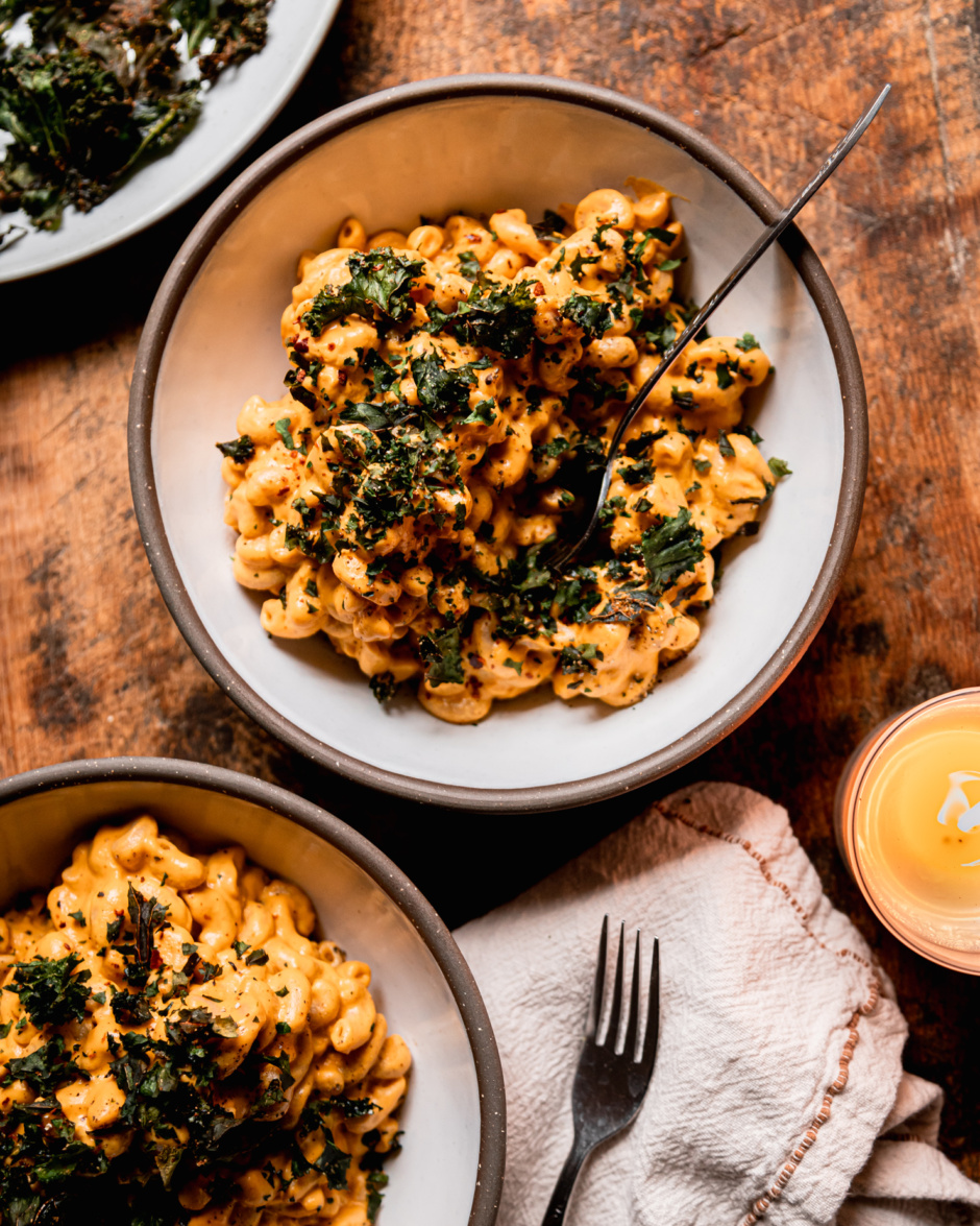 An overhead shot shows a bowl of creamy truffle mac pasta topped with crushed up bits of crunchy baked kale. A fork is sticking out of the individual serving. Another serving of the truffle mac is in the corner of the frame and a lit candle is nearby; all on a worn wood background.