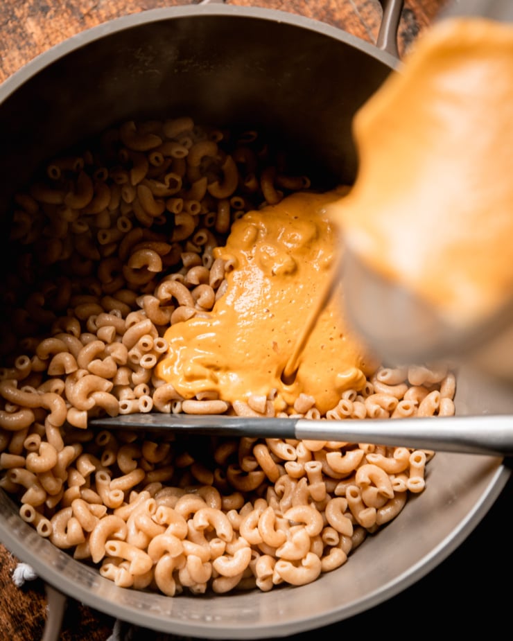 An overhead shot shows a creamy vegan truffle sauce being poured onto cooked whole wheat elbow macaroni in a pot.