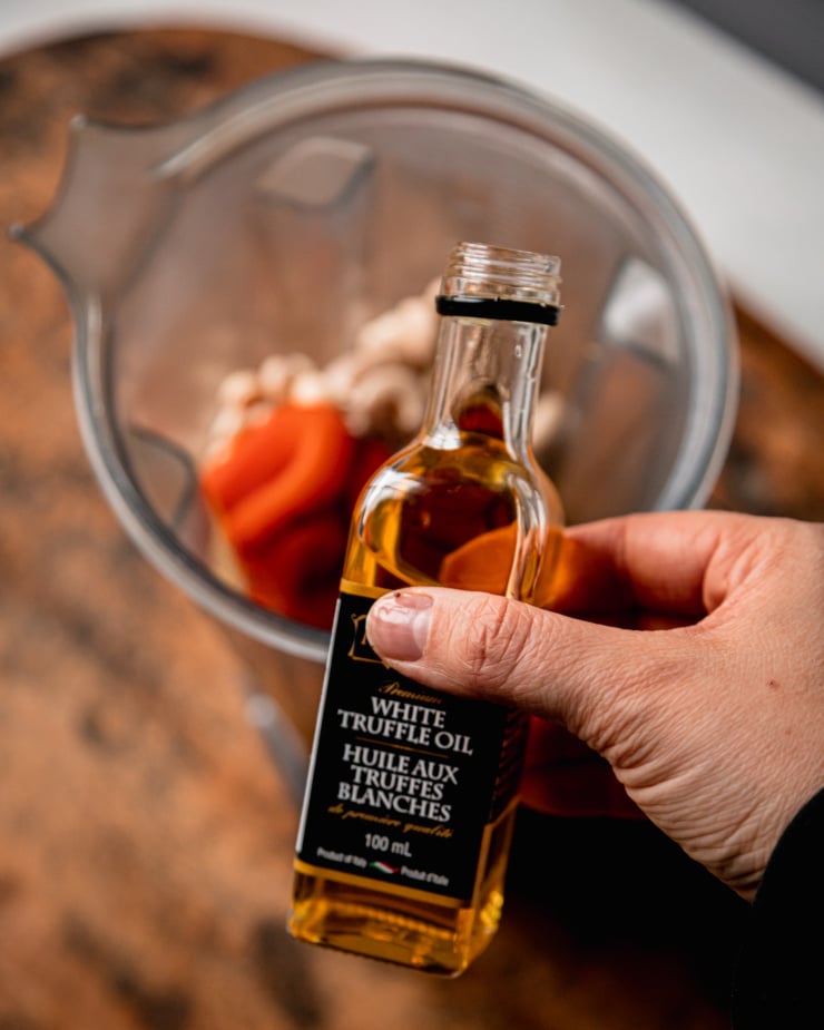 An overhead shot shows a hand holding a small bottle of truffle oil near a blender pitcher.
