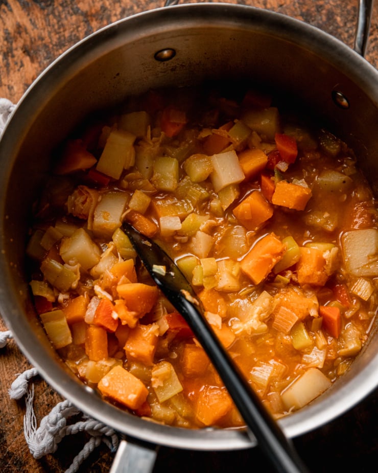 An overhead shot shows a pot of mixed cooked diced vegetables with a bit of broth. A spatula is sticking out of the pot.