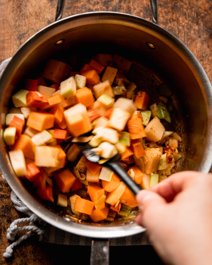 An overhead shot shows diced vegetables in a pot being stirred up by a hand holding a spatula.