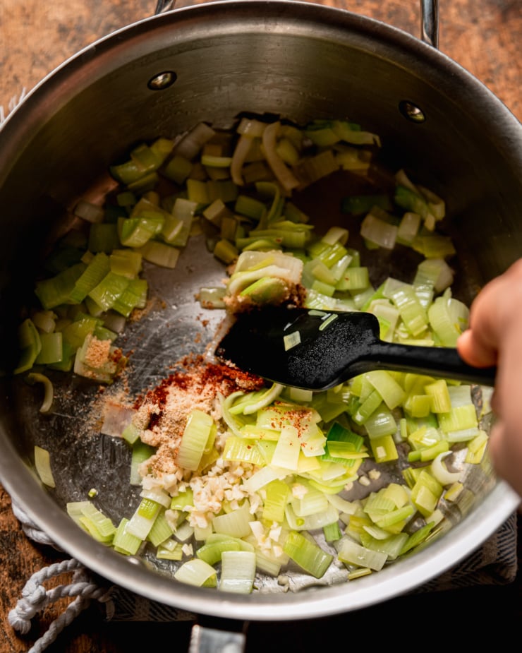 An overhead shot shows sliced leeks being sautéed in a medium stainless steel saucepan with garlic and some spices.