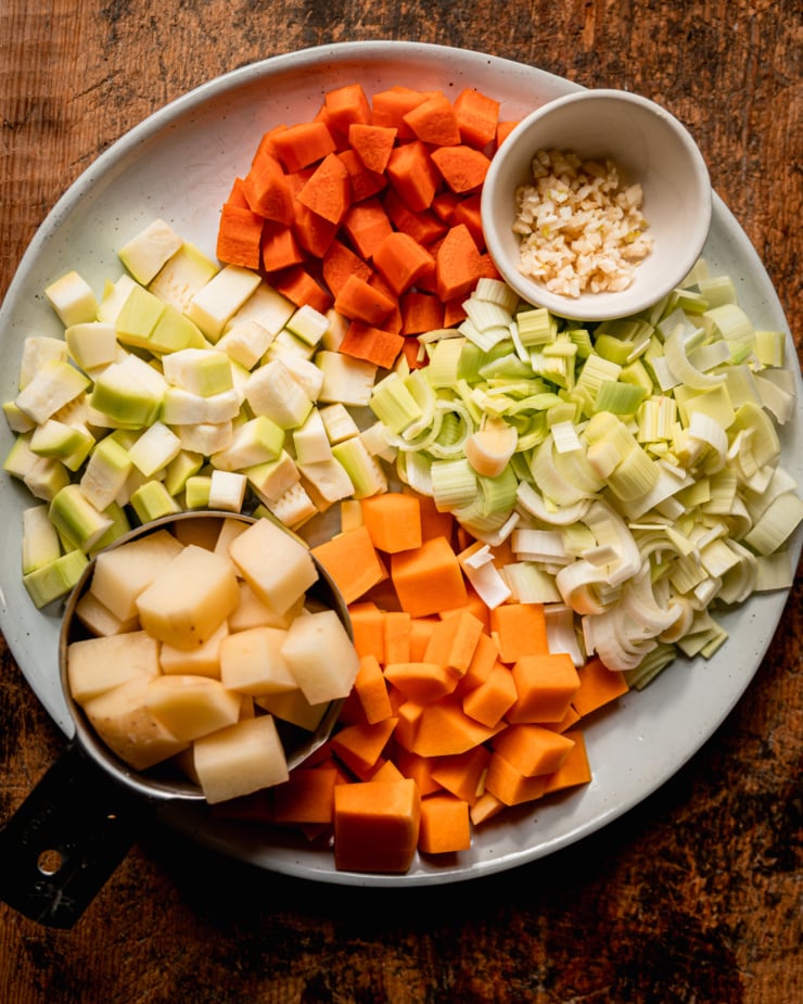 An overhead shot shows vegetable prep for a creamy truffle mac sauce: diced carrots, minced garlic, sliced leeks, diced butternut squash, diced potato in a measuring cup, and diced peeled zucchini; all on a plate.