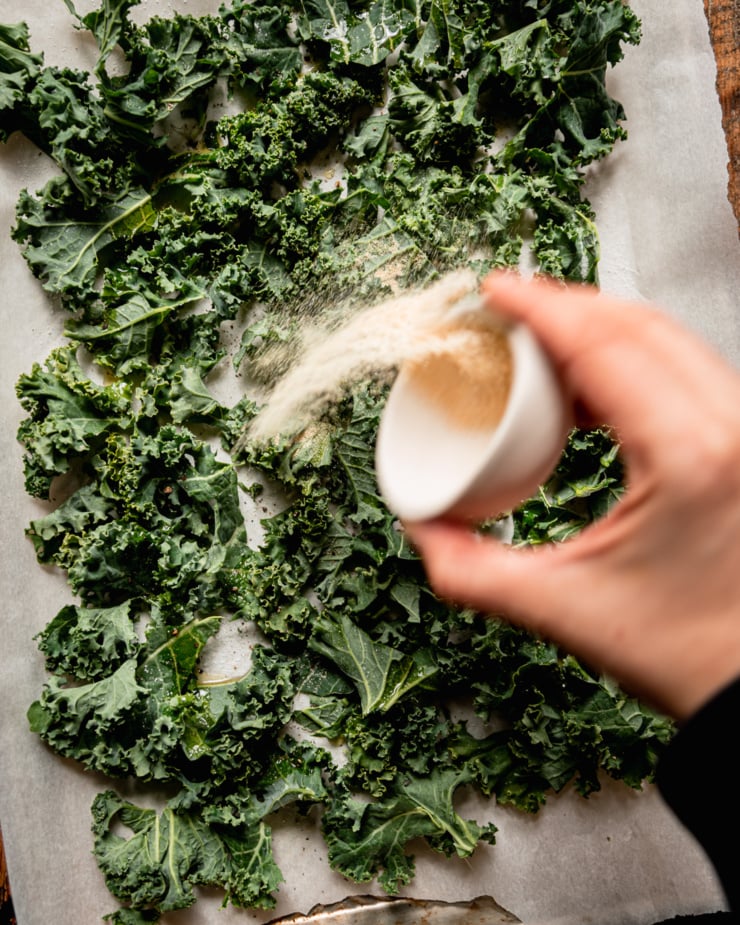An overhead shot shows a hand emptying a small bowl of spices onto chopped kale on a baking sheet.