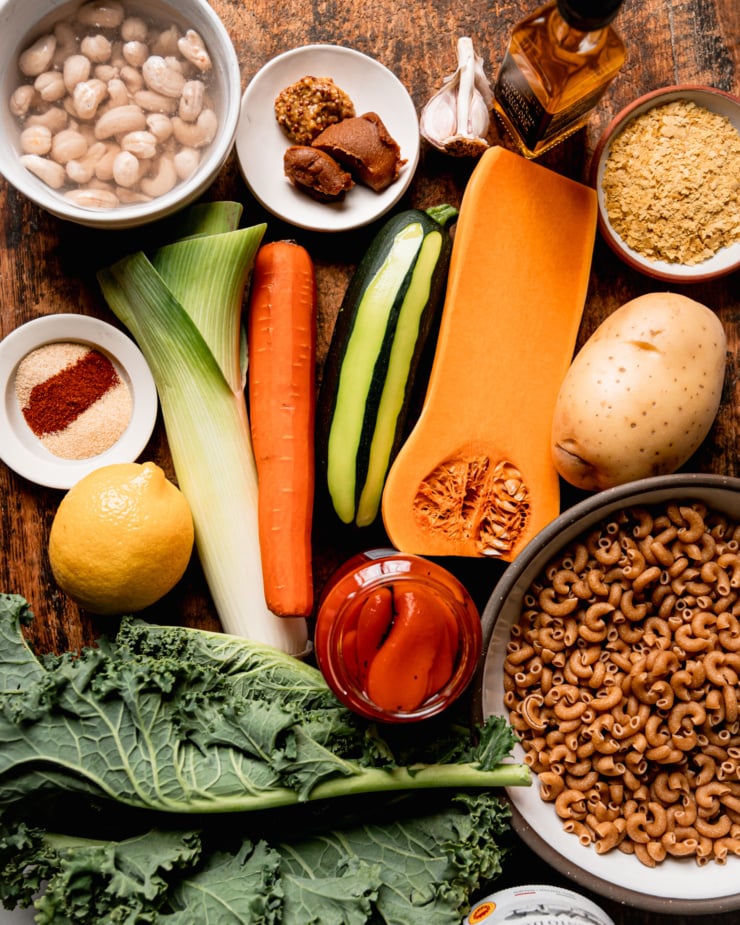 An overhead shot shows ingredients for a creamy vegan truffle mac pasta dish: miso, grainy mustard, garlic, truffle oil, nutritional yeast, butternut squash, potato, whole wheat pasta, roasted red peppers in a jar, kale, zucchini, carrot, leek, lemon, spices, and raw cashews soaking in water.