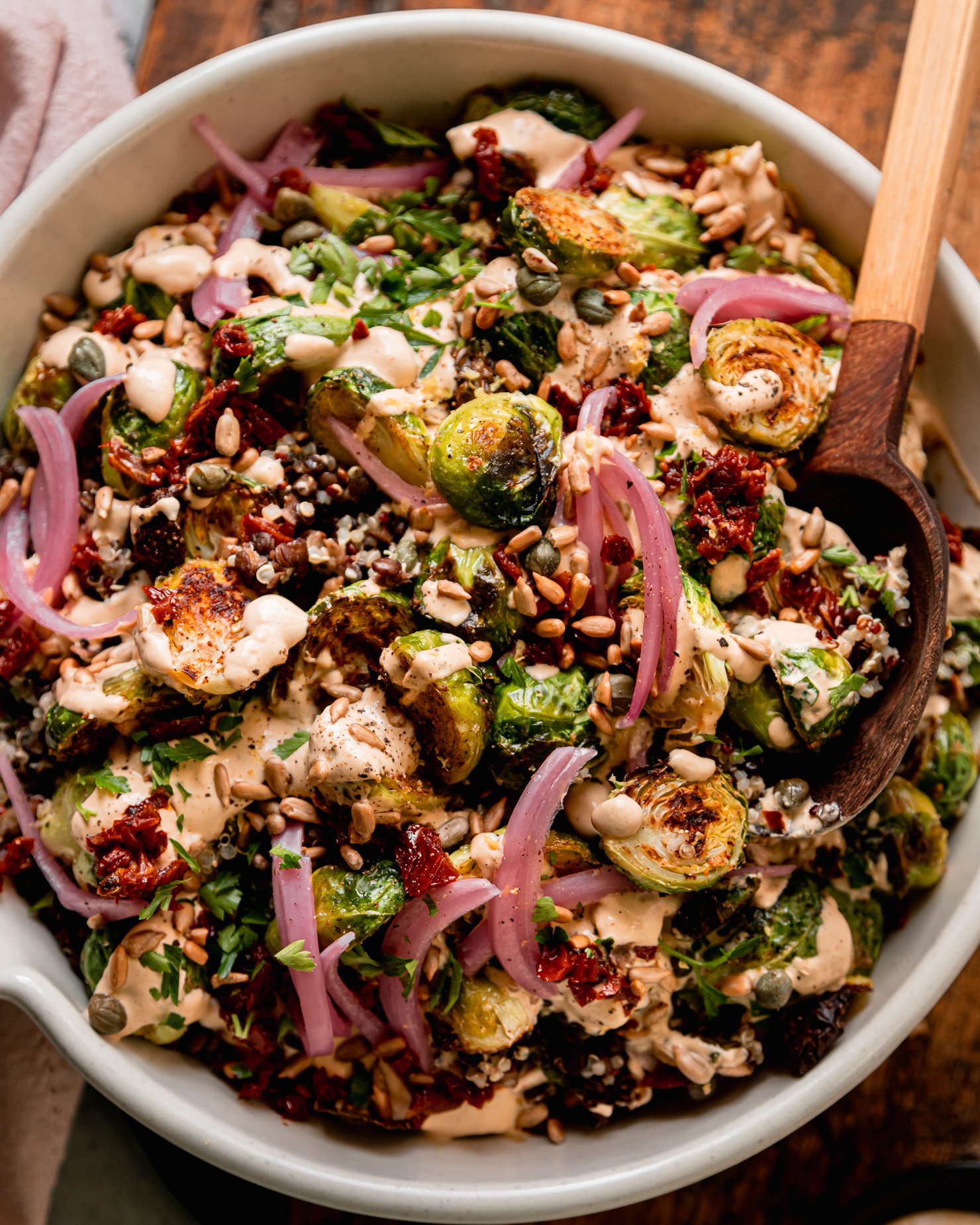 An up close, overhead shot shows a bowl filled with a roasted brussels sprouts, quinoa, and lentil salad with spicy caesar dressing drizzled over the top. The salad is garnished with chopped sun dried tomatoes, pickled red onions, toasted sunflower seeds and chopped parsley. A wooden serving spoon is sticking out of the bowl.