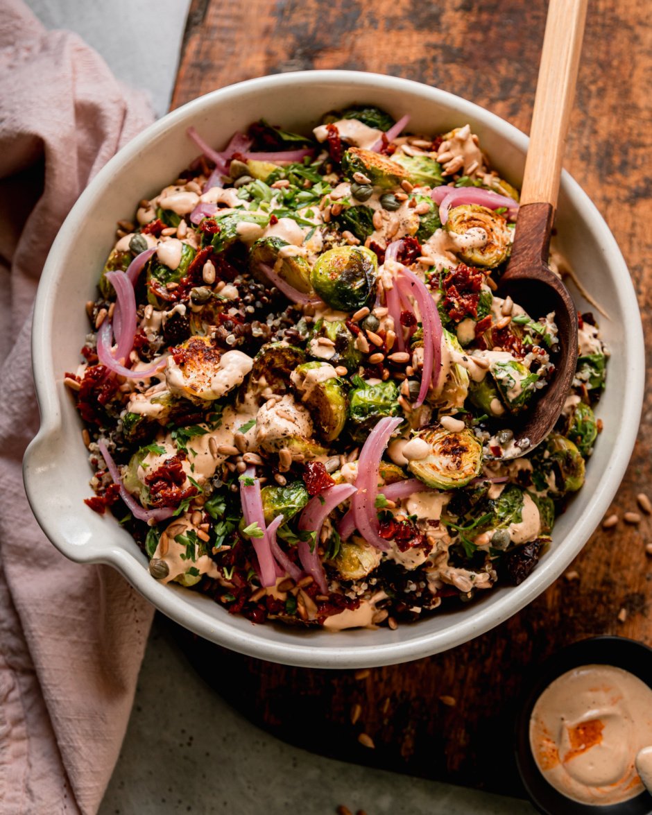 An overhead shot shows a bowl filled with a roasted brussels sprouts, quinoa, and lentil salad with spicy caesar dressing drizzled over the top. The salad is garnished with chopped sun dried tomatoes, pickled red onions, toasted sunflower seeds and chopped parsley. A pink napkin is nearby and the bowl is perched on a rough wooden board.