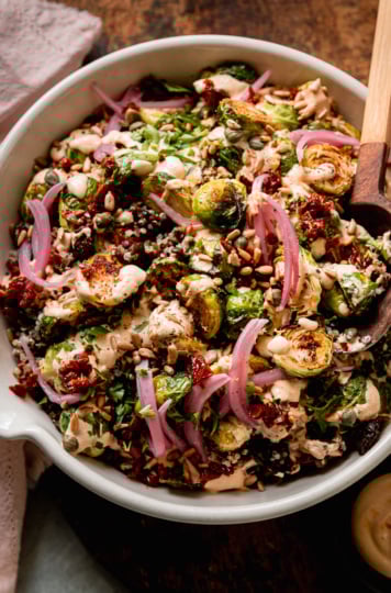 An overhead shot shows a bowl filled with a roasted brussels sprouts, quinoa, and lentil salad with spicy caesar dressing drizzled over the top. The salad is garnished with chopped sun dried tomatoes, pickled red onions, toasted sunflower seeds and chopped parsley. A pink napkin is nearby.