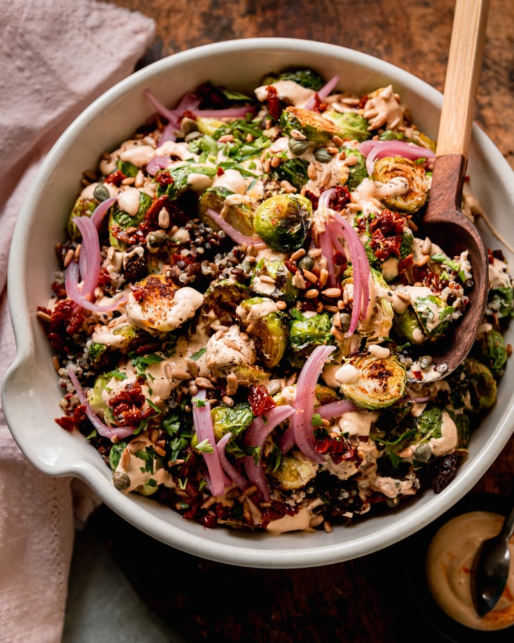 An overhead shot shows a bowl filled with a roasted brussels sprouts, quinoa, and lentil salad with spicy caesar dressing drizzled over the top. The salad is garnished with chopped sun dried tomatoes, pickled red onions, toasted sunflower seeds and chopped parsley. A pink napkin is nearby.