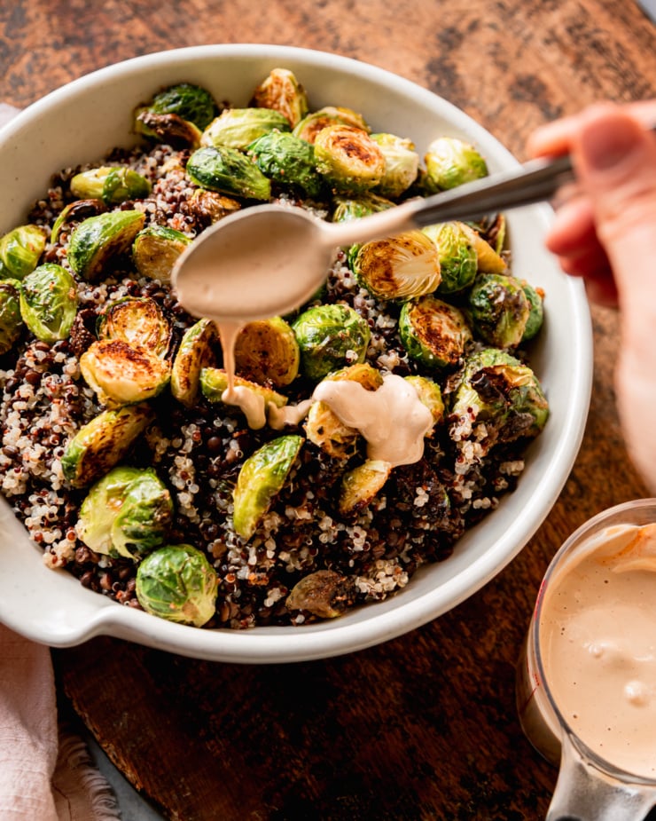 A 3/4 angle shot shows a hand using a spoon to drizzle a creamy dressing over a bowl with cooked quinoa, lentils, and roasted brussels sprouts.