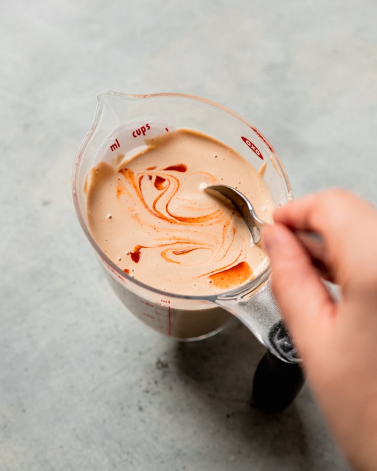 An overhead shot shows a hand using a spoon to stir some hot sauce into a measuring cup filled with a creamy dressing.