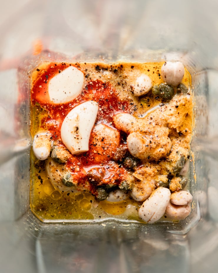 An up close, overhead shot shows a view into a blender jar with ingredients for a spicy vegan caesar dressing.