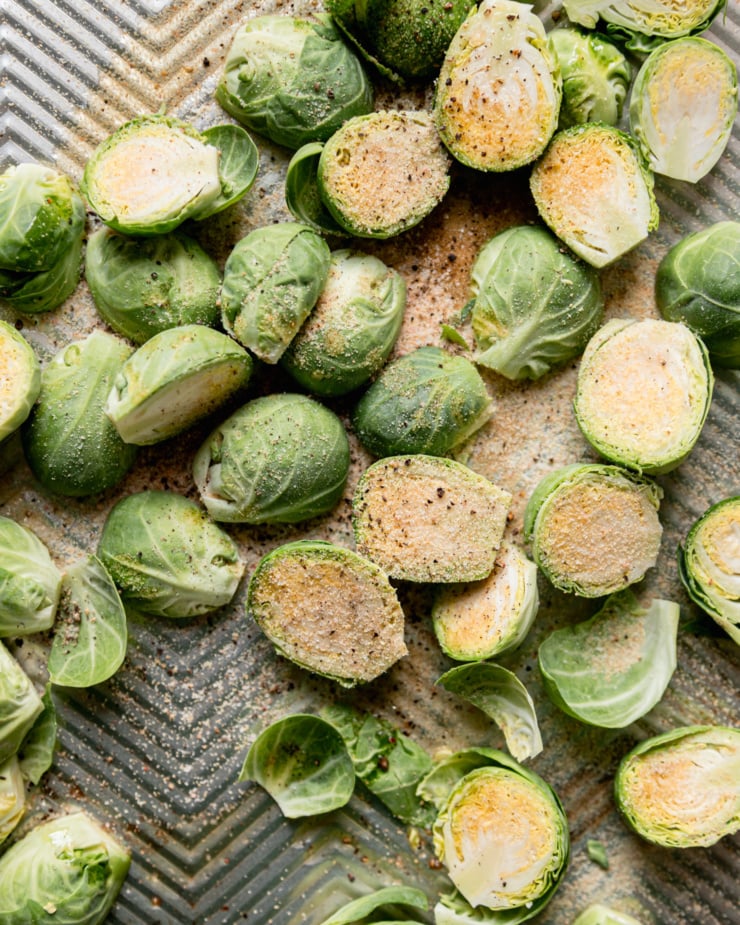 An up close, overhead shot shows halved brussels sprouts on a baking sheet dusted with spices, salt, and pepper.