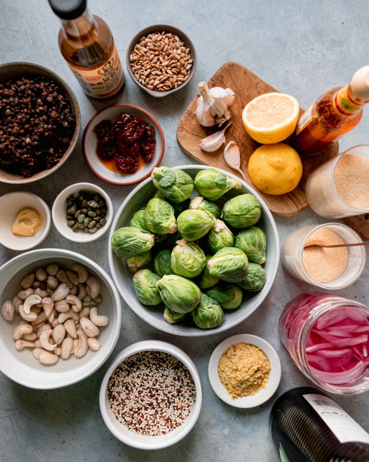 An overhead shot shows ingredients used in a Brussels sprouts, quinoa and lentil salad with spicy vegan caesar dressing. Most ingredients are in bowls or jars against a blue-ish grey background.