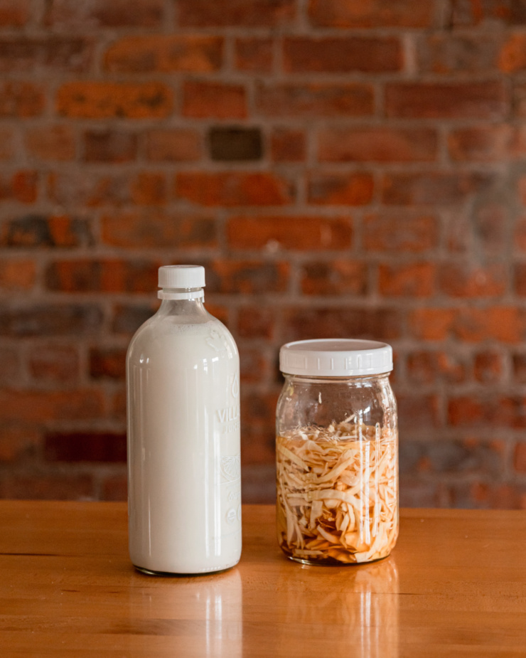 A head-on shot shows a bottle of homemade nutmeg and a jar of pickled cabbage on a counter against a brick background.