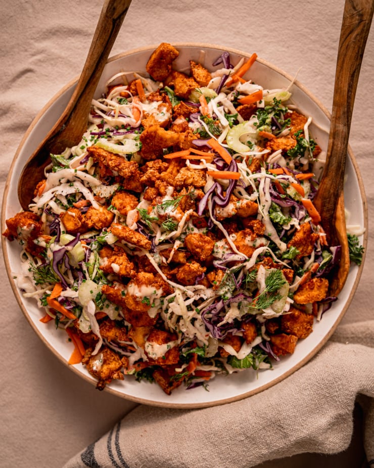 An overhead shot shows a wide serving bowl filled with vegan ranch slaw and topped with crunchy baked Buffalo tofu pieces. A set of wooden tongs is perched int he slaw as well.