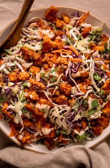 An up close, overhead shot shows a wide serving bowl filled with vegan ranch slaw and topped with crunchy baked Buffalo tofu pieces. A set of wooden tongs is perched int he slaw as well.