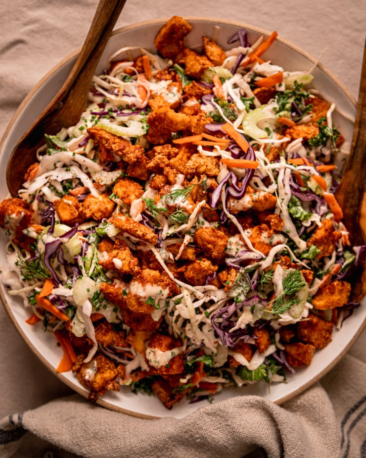 An up close, overhead shot shows a wide serving bowl filled with vegan ranch slaw and topped with crunchy baked Buffalo tofu pieces. A set of wooden tongs is perched int he slaw as well.
