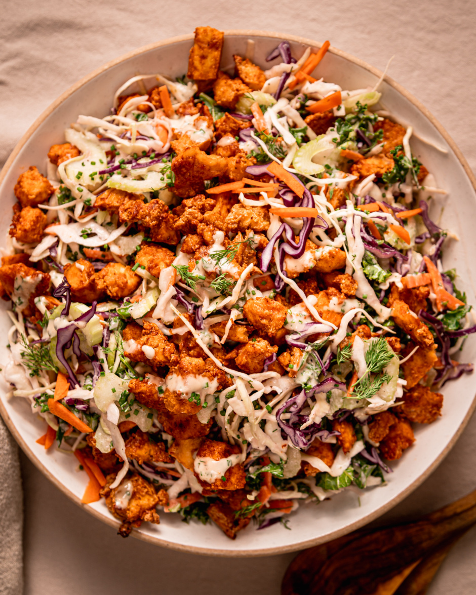 An up close, overhead shot shows a wide serving bowl filled with colourful vegan ranch slaw that's topped with crunchy baked Buffalo tofu pieces.