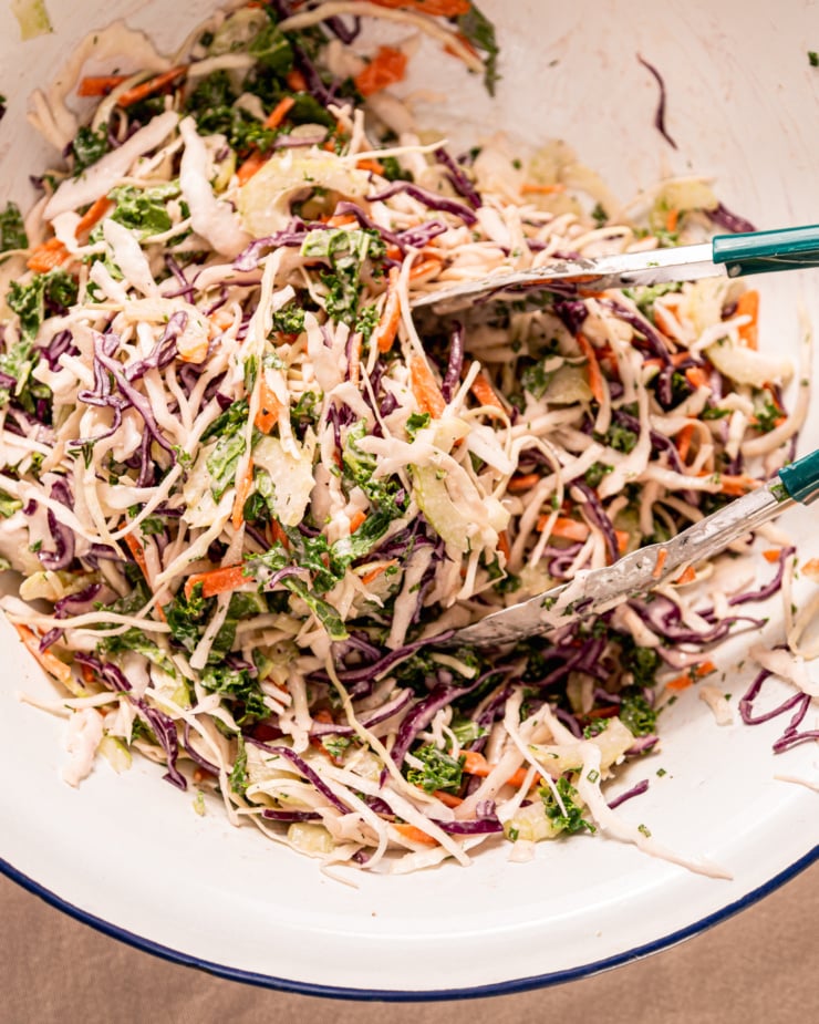 An overhead shot shows a colourful and creamy vegan ranch slaw being tossed up in a bowl with some tongs.