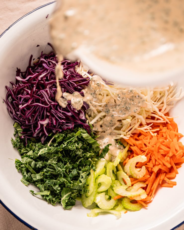 An overhead shot shows a creamy dressing being poured over shredded cabbage, carrots, sliced kale, and chopped celery.